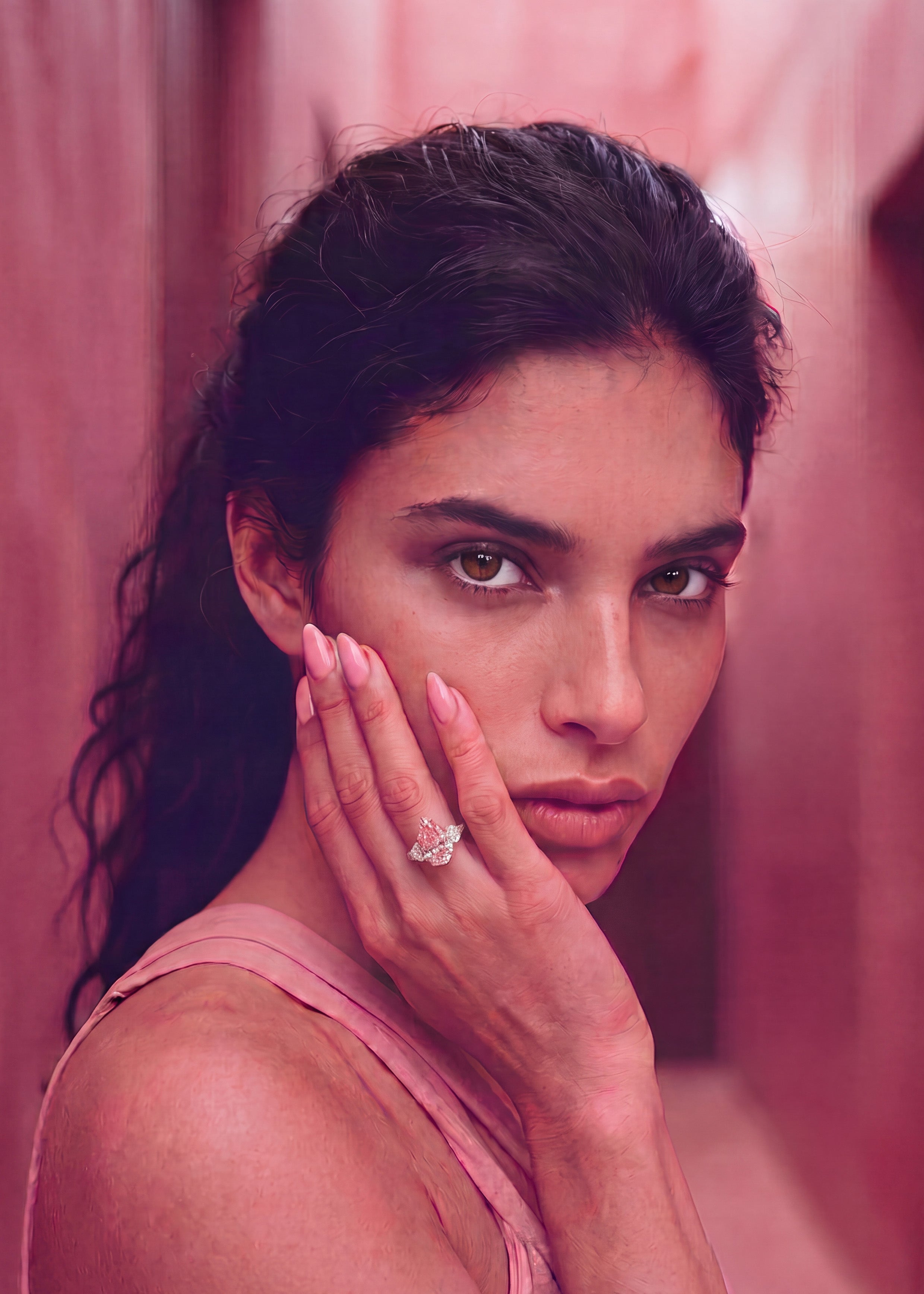 A close-up portrait of a woman with dark hair, wearing a pink top and holding her hand to her face, showcasing a ring with a large clear gemstone on her finger. The background is a soft, draped pink fabric.