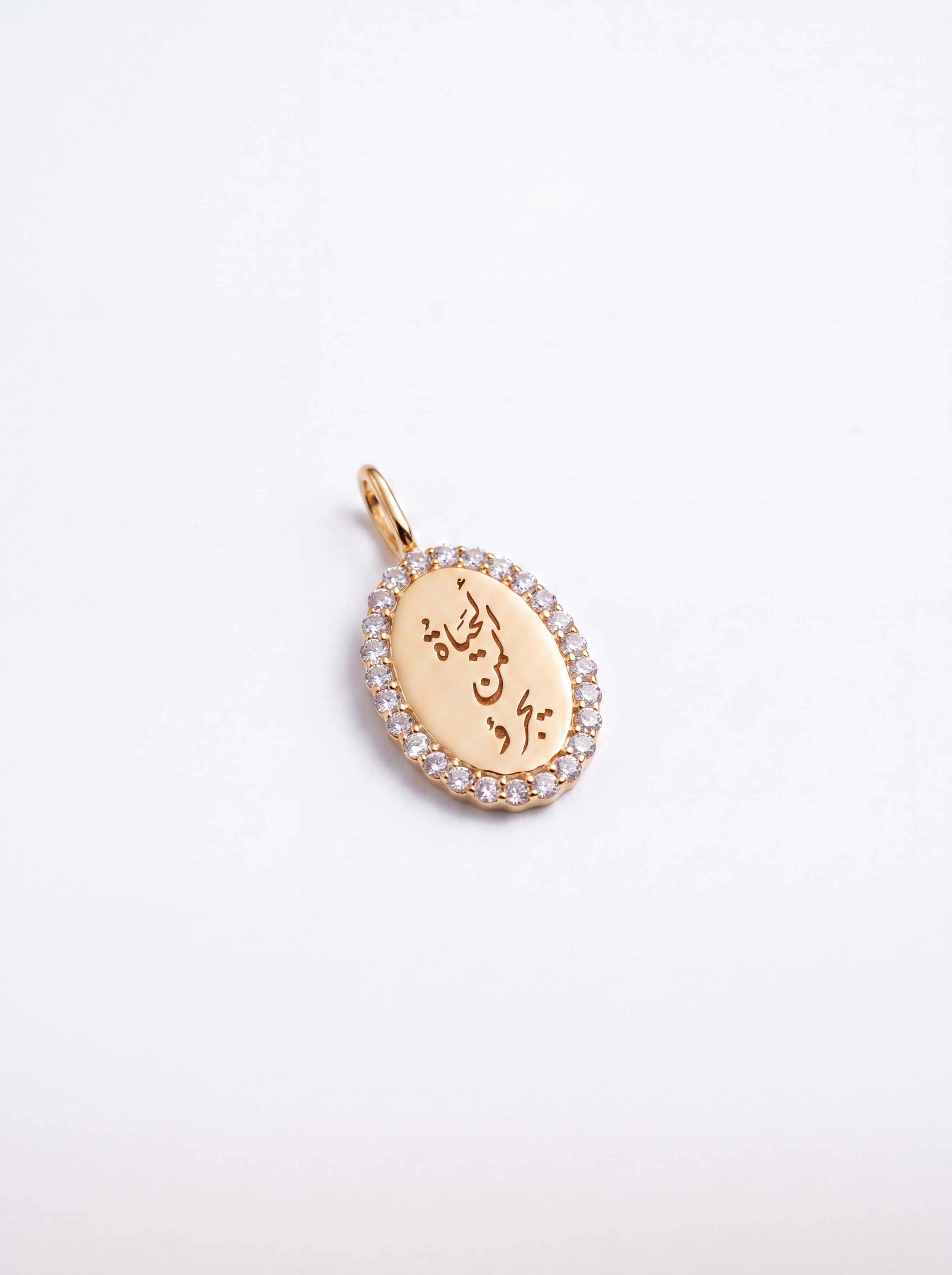 An oval gold pendant featuring engraved Arabic calligraphy and a border of small clear stones, photographed on a clean white background.