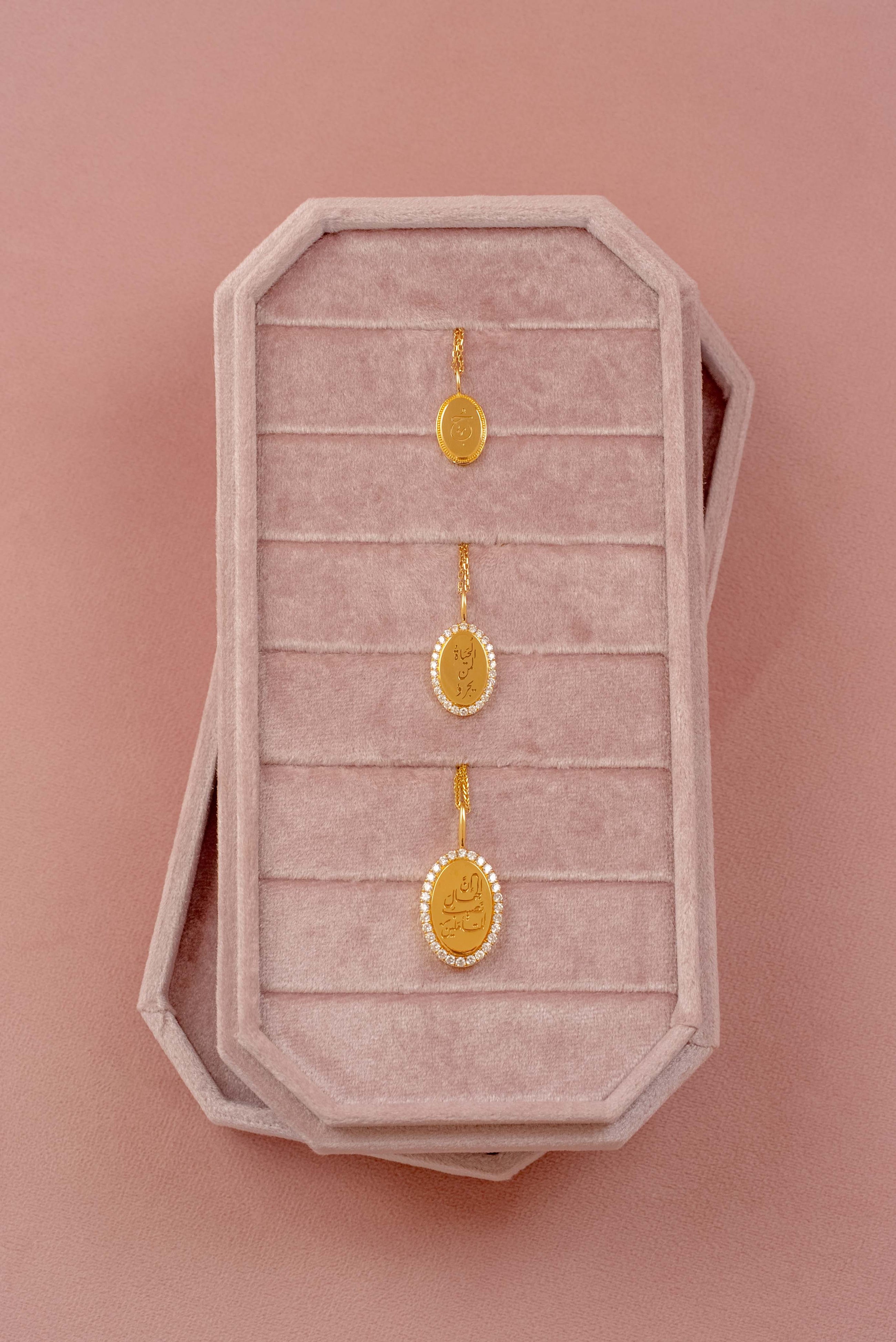 Three gold oval pendants displayed on a pink velvet jewelry box against a solid pink background.