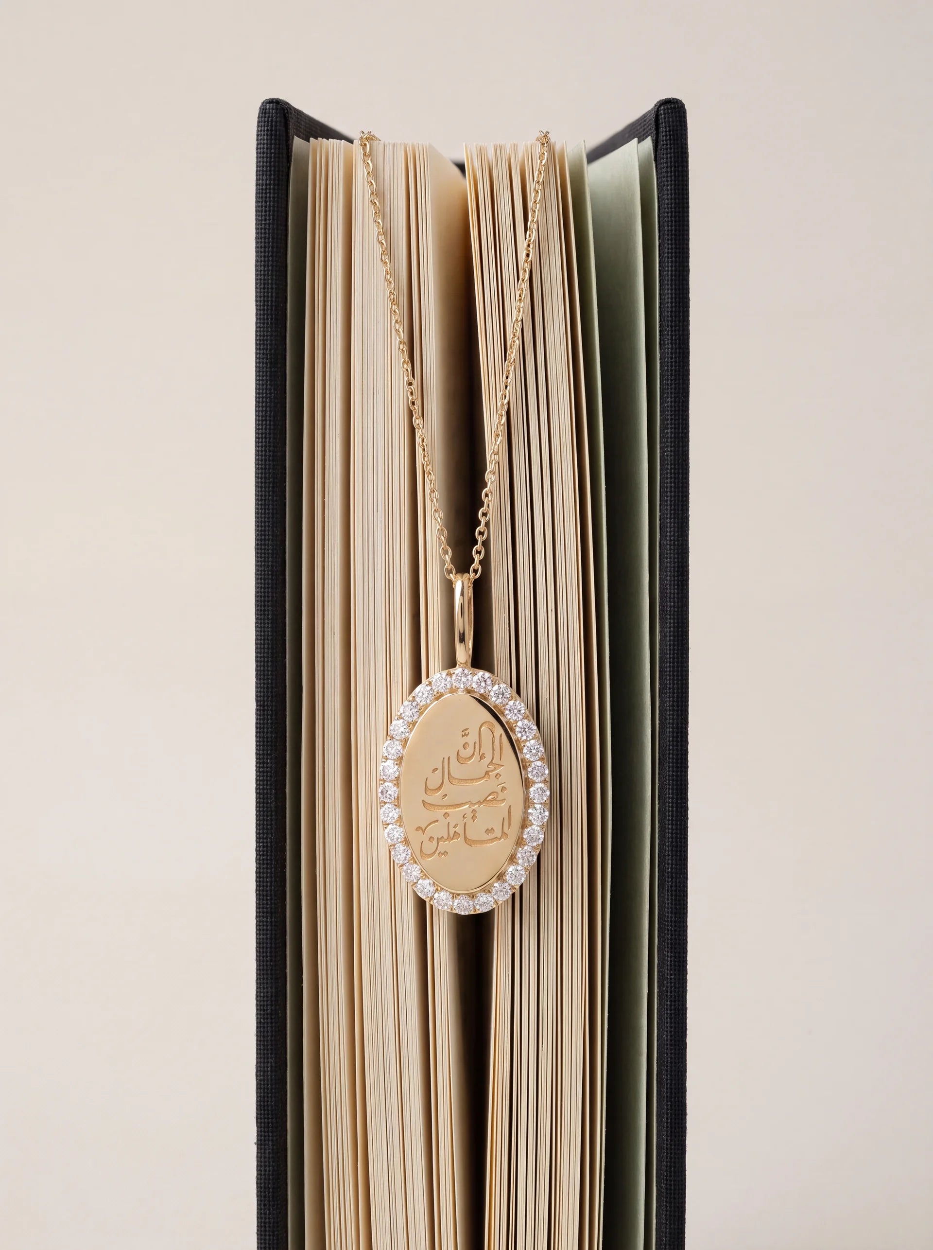 A gold oval pendant necklace with a border of diamonds and Arabic calligraphy in the center, draped over the open pages of a black book against a light beige background.