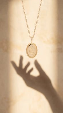 A gold oval pendant necklace with a textured border and a central Arabic calligraphy, hanging from a delicate gold chain. The pendant is shown against a warm, sunlit beige background with a prominent hand shadow cast below.