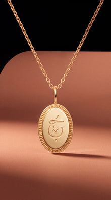 A gold oval pendant necklace with an engraved symbol, displayed on a warm peach-colored curved surface against a dark background.