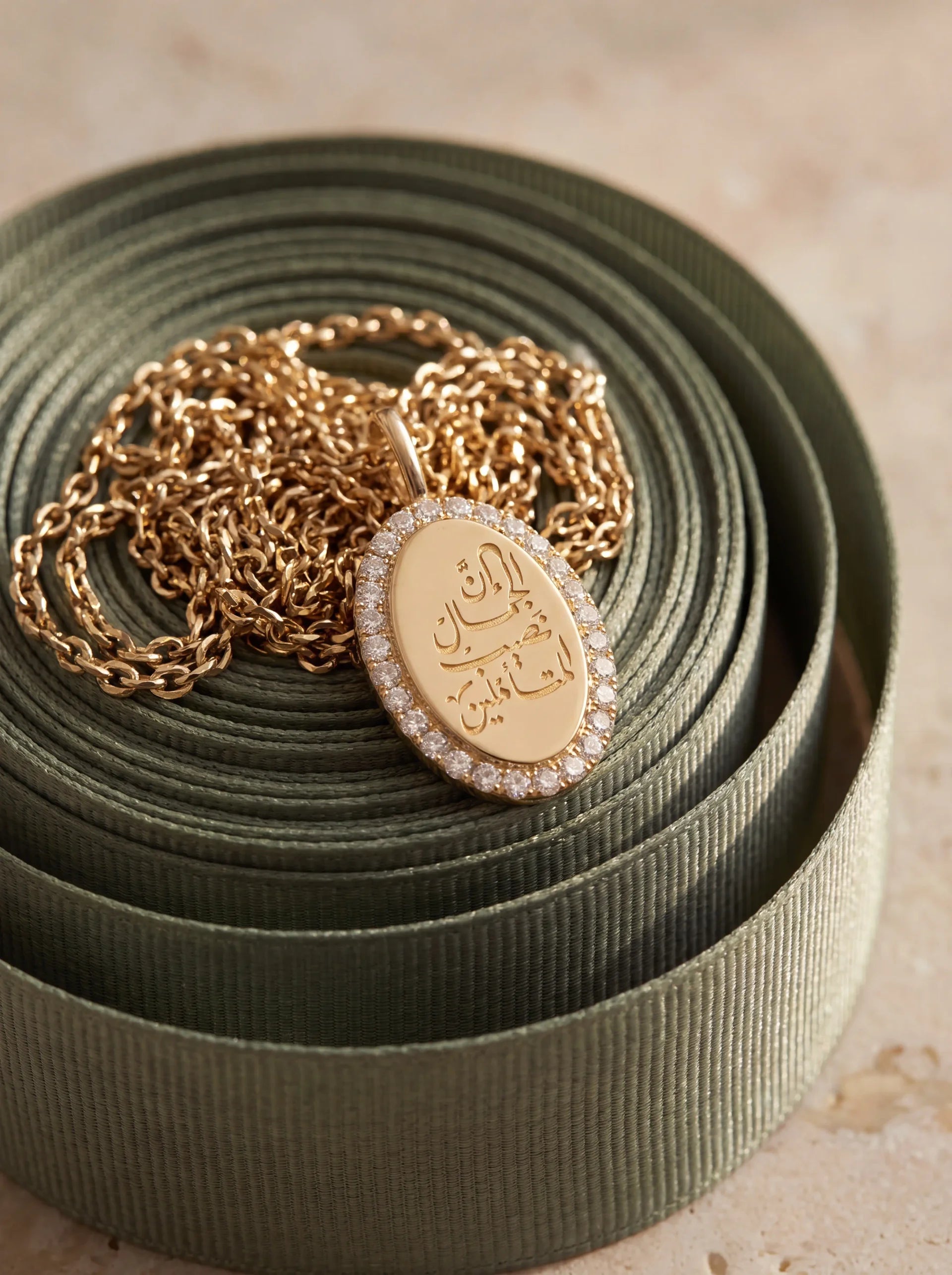 A gold oval pendant necklace with Arabic calligraphy engraved on the front, surrounded by a halo of small clear stones, resting on a coiled olive green ribbon over a neutral beige background.