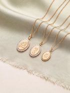 Three gold-toned oval pendant necklaces with engraved script and small clear stones, arranged on a soft cream satin fabric background.