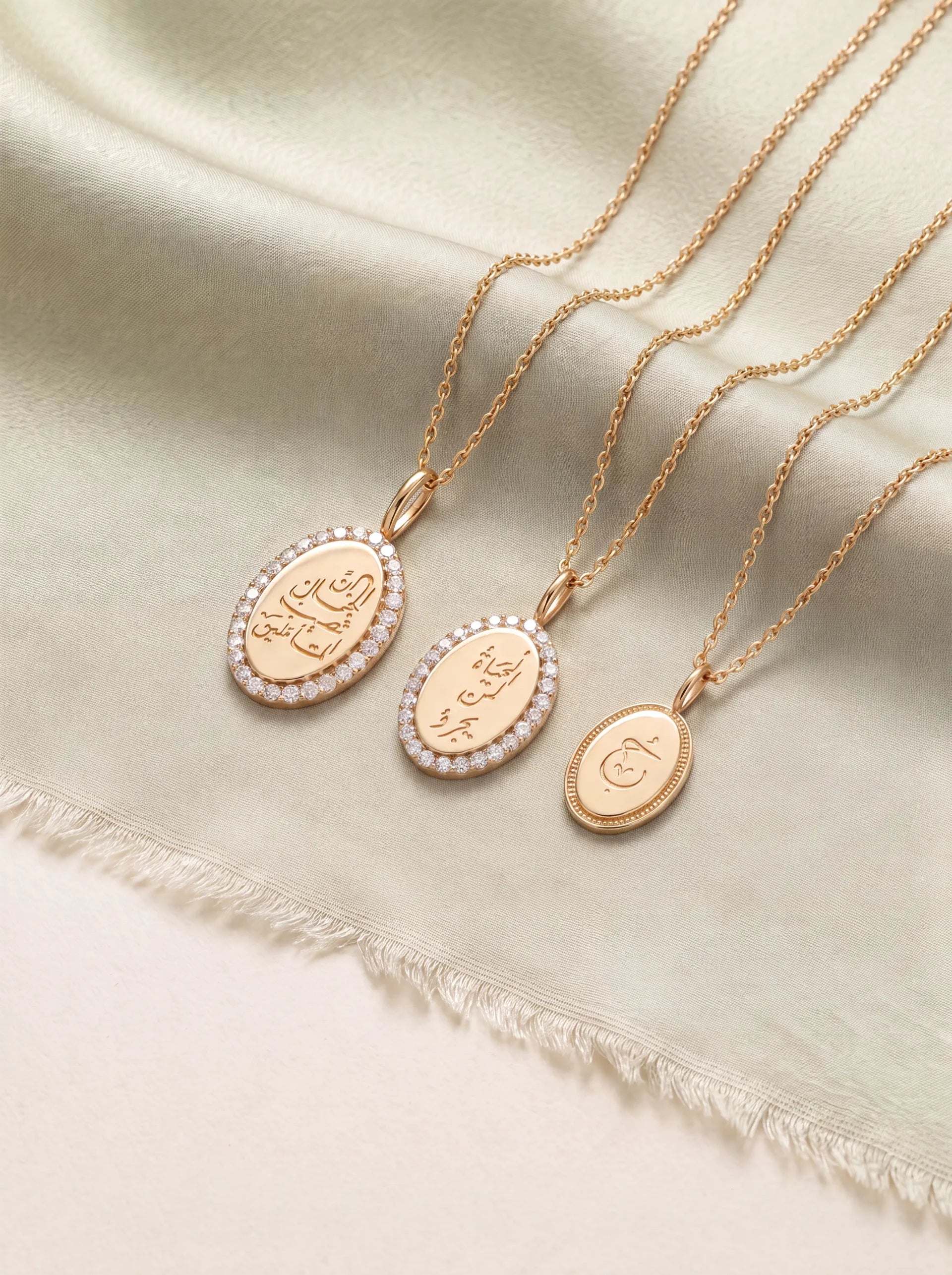 Three gold-toned oval pendant necklaces with engraved script and small clear stones, arranged on a soft cream satin fabric background.
