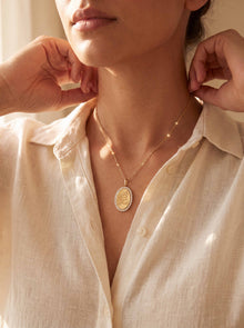 A close-up lifestyle shot of a model wearing a delicate gold necklace with an oval pendant featuring an embossed profile and a border of small clear stones, styled with a matching beaded chain.