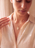 A close-up lifestyle shot of a woman wearing a delicate gold necklace with an oval pendant, styled with a light beige linen button-up shirt.
