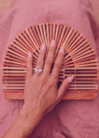 A close-up of a hand resting on a semi-circular wooden slat clutch bag, with a pink pear-shaped  ring worn on one finger, set against a dusty pink fabric background.