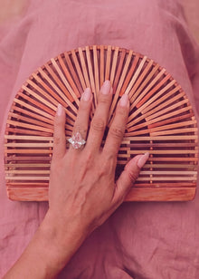 A close-up of a hand resting on a semi-circular wooden slat clutch bag, with a pink pear-shaped  ring worn on one finger, set against a dusty pink fabric background.