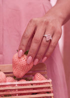 A close-up lifestyle shot of a woman's hand with pink manicured nails wearing a pink bow ring, holding a pink strawberry over a wooden crate of strawberries.