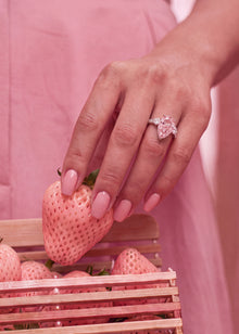 A close-up lifestyle shot of a woman's hand with pink manicured nails wearing a pink bow ring, holding a pink strawberry over a wooden crate of strawberries.