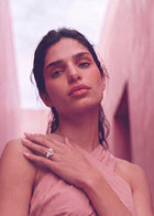 A close-up portrait of a woman wearing a pink draped dress and a prominent diamond ring on her left hand, set against a soft pink background.