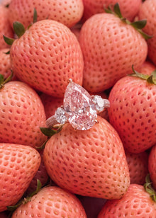 A pear-cut diamond ring with round diamonds on a silver band, resting on top of a pile of fresh pinkish-red strawberries.