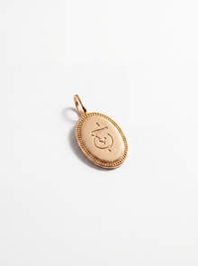 A small oval gold pendant with a decorative border and an engraved symbol in the center, shown on a clean white background.