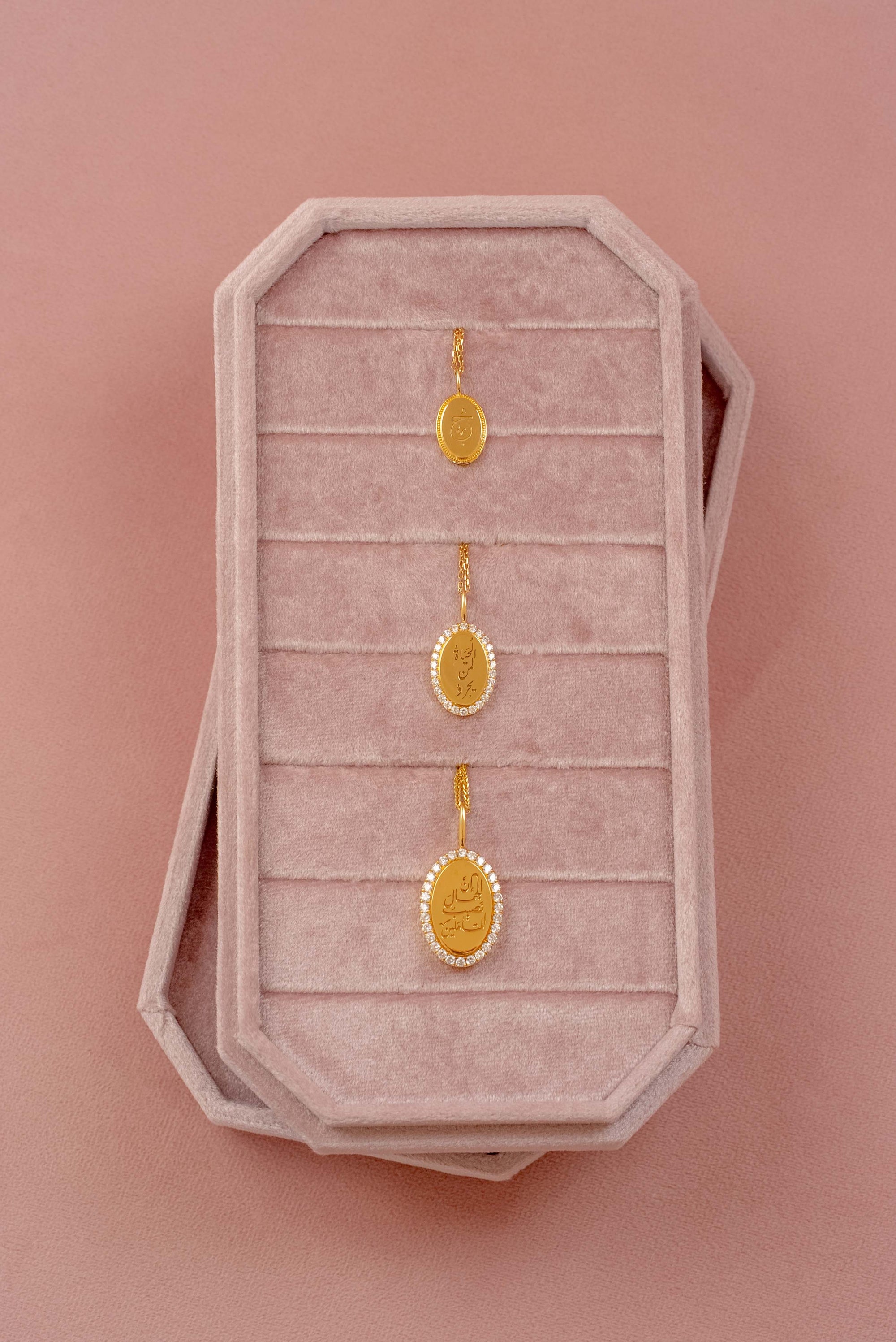 Three gold oval pendants displayed on a pink velvet jewelry box against a solid pink background.