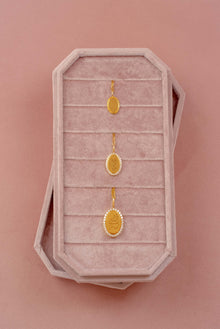 Three gold oval pendants displayed on a pink velvet jewelry box against a solid pink background.