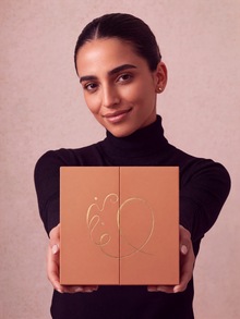 A woman in a black turtleneck sweater holds up a square peach-colored box with a gold foil monogram logo, set against a soft pink textured background.