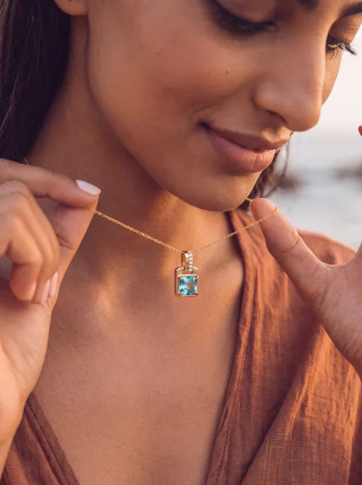 A close-up lifestyle shot of a woman wearing a gold necklace with a square-shaped light blue gemstone pendant, holding the chain with both hands against a warm, blurred background.