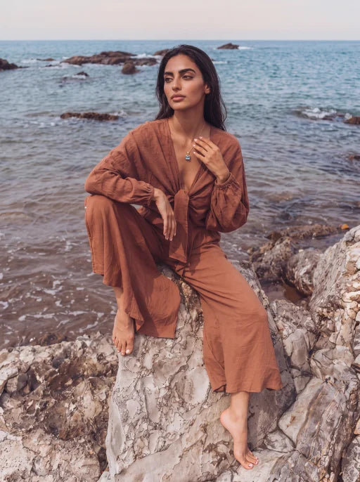 A woman wearing a rust-brown long-sleeve wrap-style jumpsuit sits barefoot on a rocky shoreline with the ocean in the background.