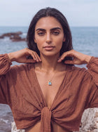 A woman with dark hair stands on a rocky beach with the ocean in the background, wearing a rust-brown tie-front top and a delicate gold necklace with a light blue pendant.