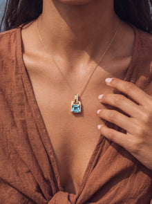 A close-up lifestyle shot of a model wearing a gold chain necklace with a square-cut light blue gemstone pendant, styled with a brown linen top.