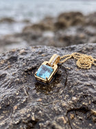 A gold pendant necklace with a rectangular light blue gemstone and a small pave accent on the bail, resting on a wet dark rock with a blurred ocean background.