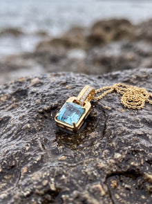 A gold pendant necklace with a rectangular light blue gemstone and a small pave accent on the bail, resting on a wet dark rock with a blurred ocean background.