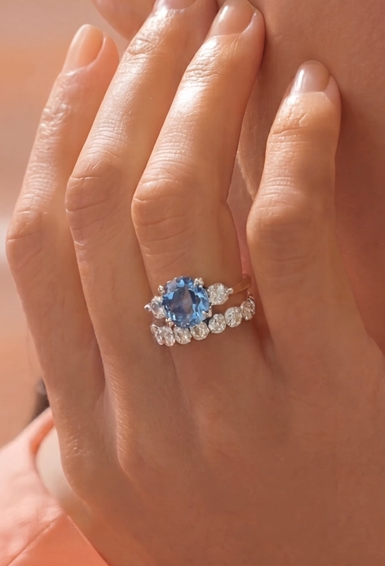 Close-up of a hand wearing a blue diamond ring with a white diamond band on a soft pink background