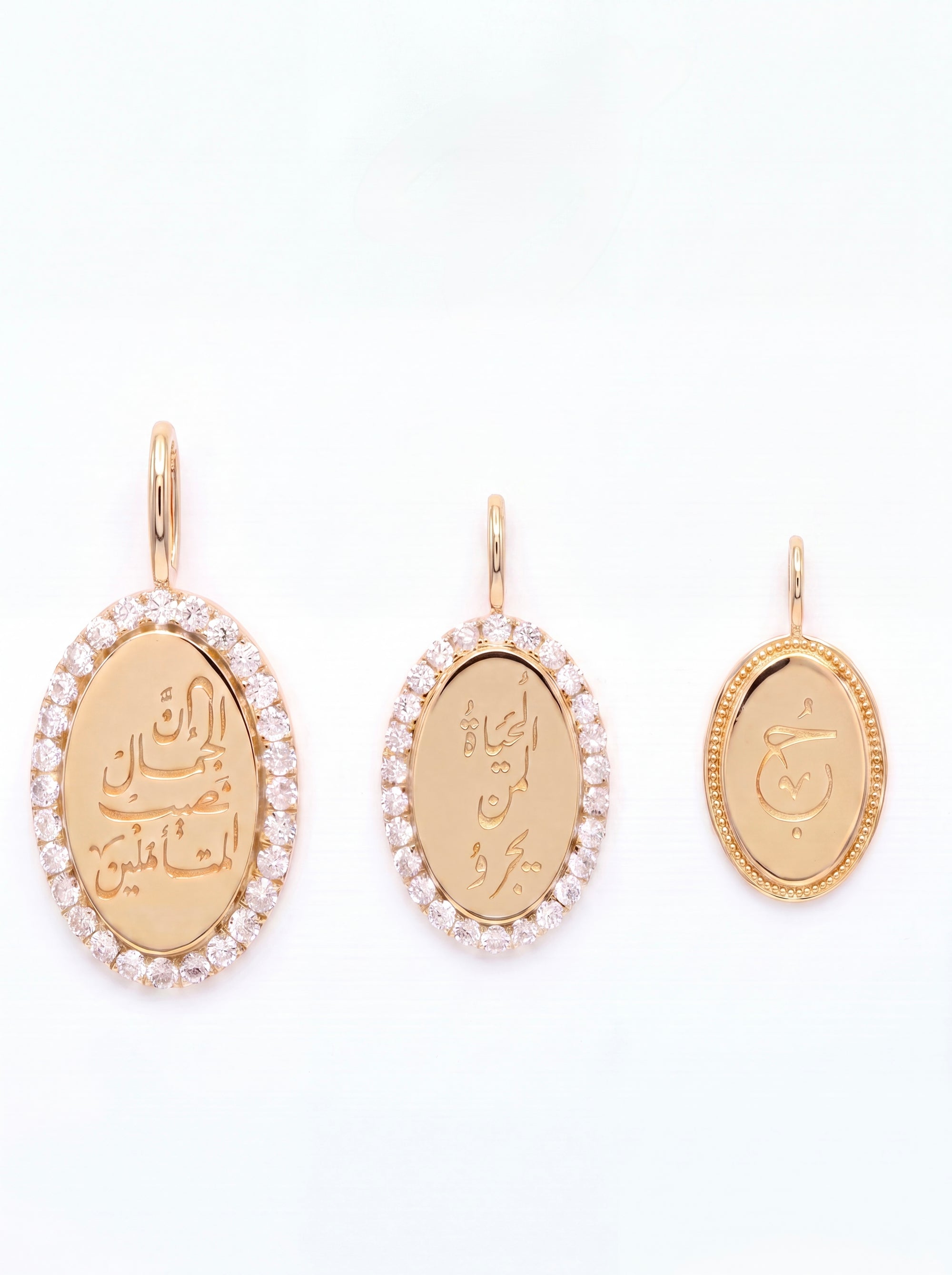 Three gold oval pendant charms with Arabic calligraphy, displayed in three different sizes on a plain white background. The two larger pendants are surrounded by a halo of small clear stones, and the smallest pendant on the right has a textured gold border.