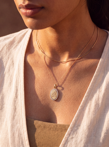A close-up lifestyle shot of a model wearing a gold double-layer necklace with an oval pendant featuring a white floral border and a central gold monogram.