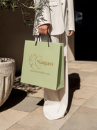 A person in a white blazer and wide-leg trousers holds a light green Manjam jewelry shopping bag with black handles, standing on a stone patio.