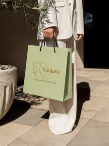 A person in a white blazer and wide-leg trousers holds a light green Manjam jewelry shopping bag with black handles, standing on a stone patio.