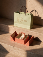 A branded Manjam paper shopping bag in pale green with dark handles sits on stone steps beside an open coral-orange jewelry box containing a ring box and ring, photographed in warm sunlight against a textured beige wall.