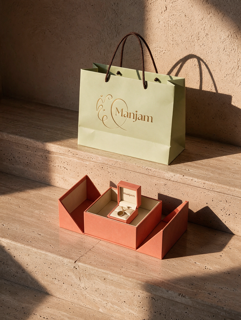 A branded Manjam paper shopping bag in pale green with dark handles sits on stone steps beside an open coral-orange jewelry box containing a ring box and ring, photographed in warm sunlight against a textured beige wall.
