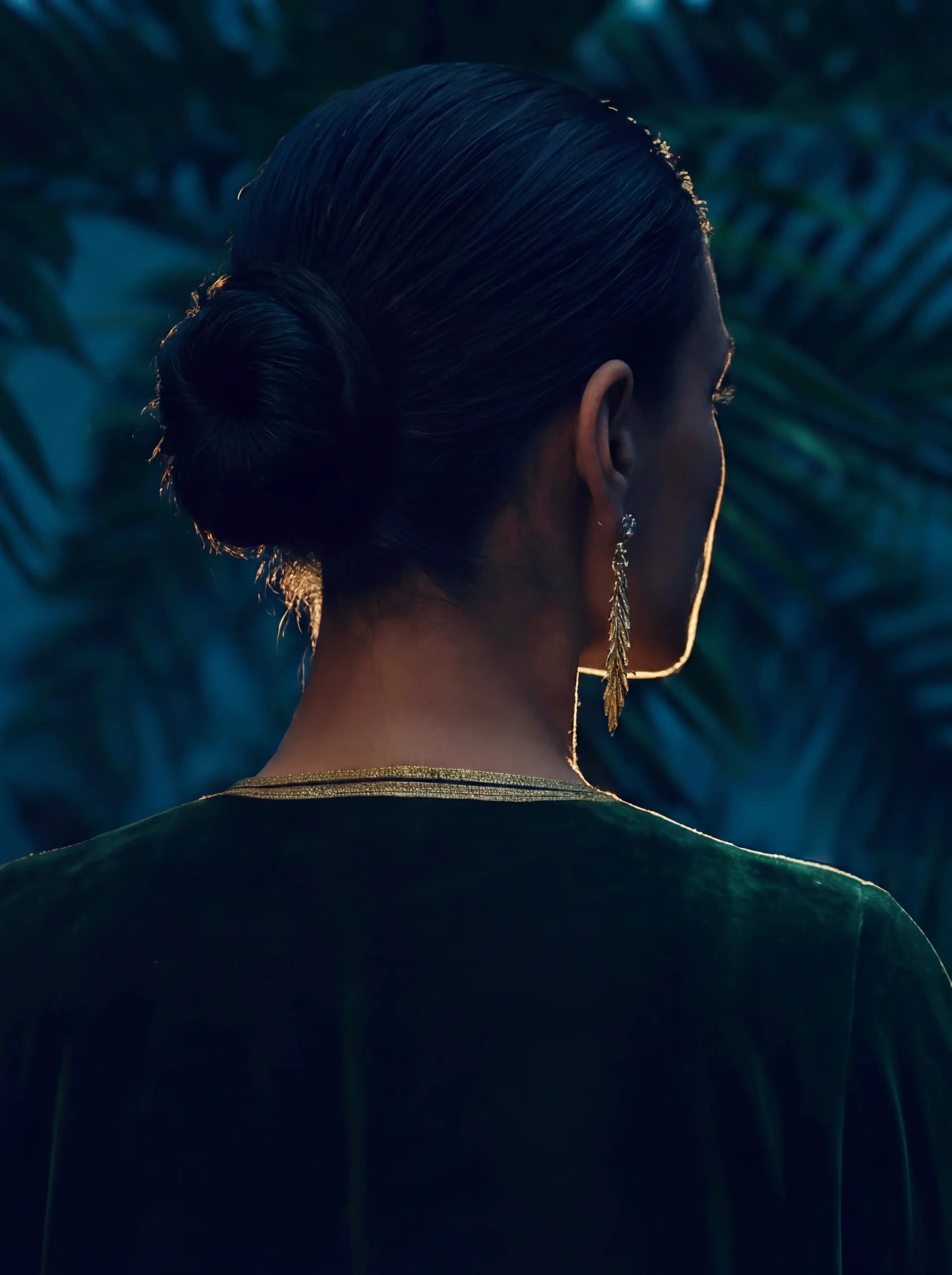 Woman with styled hair and palm leaf earrings against a dark, leafy background