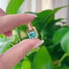 Gold pendant with a blue green gemstone held between two fingers against a green leafy background