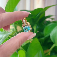 Gold pendant with a blue green gemstone held between two fingers against a green leafy background