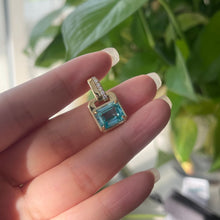 Gold earring with a blue gemstone held between fingers against a blurred green plant background