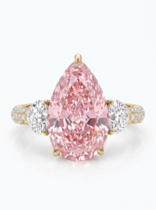 A pear-cut pink diamond ring set in yellow gold with two round diamonds on the band, photographed on a clean white background.