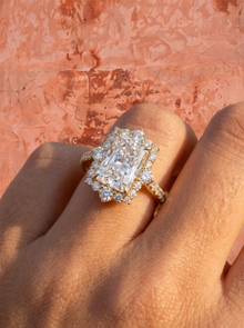 Diamond ring with a central radiant cut diamond with a diamond halo in rose gold with a large central diamond on a hand against a pink stone background